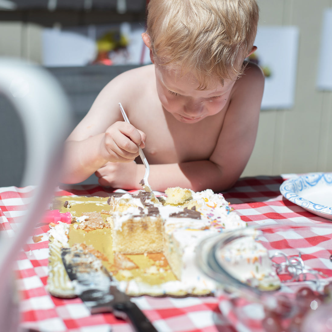 A young boy digs into a half-eaten sheet cake on a picnic table. It is an extremely sunny day, and he squints in concentration at the cake.