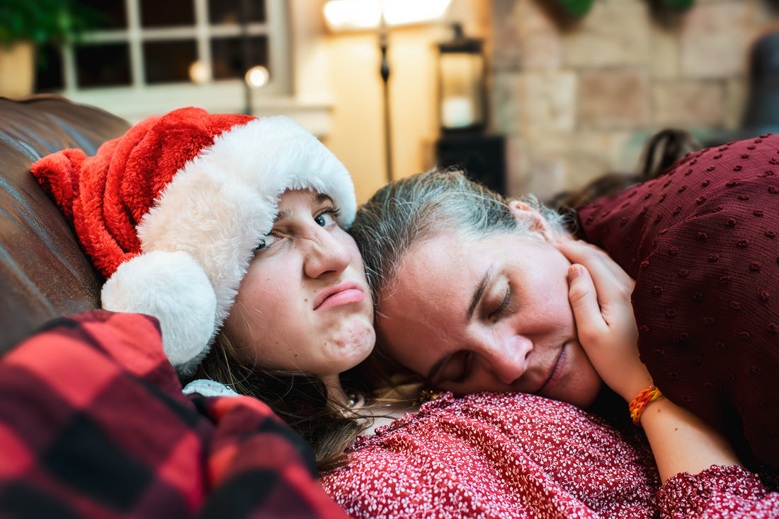 Teenage girl sitting on the couch wearing a Santa hat scowls at the camera while her mother sleeps on her shoulder.