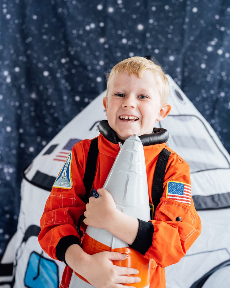 One young boy in an orange astronaut costume, hugging a rocket balloon, in front of a spaceship tent with a starry backdrop.