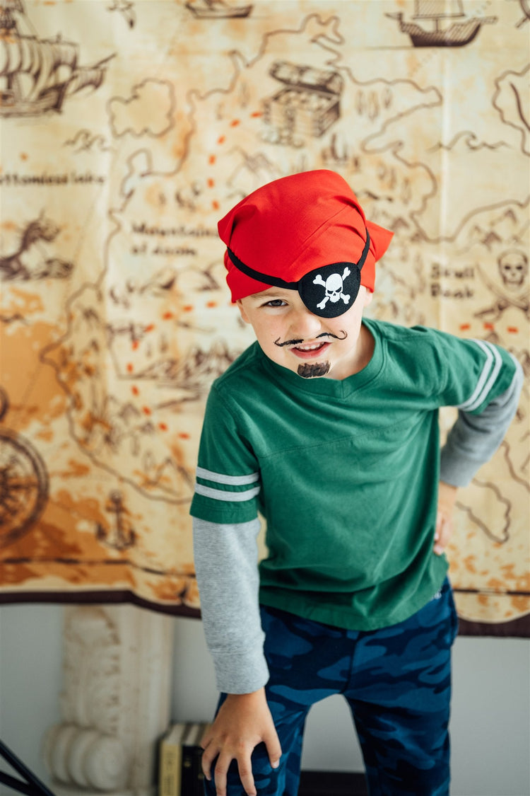 One young boy in a green shirt and jeans, leaning forward with his hand on his knee, wearing a pirate red bandana and eye patch, with a fake mustache and go tee, in front of a treasure map.